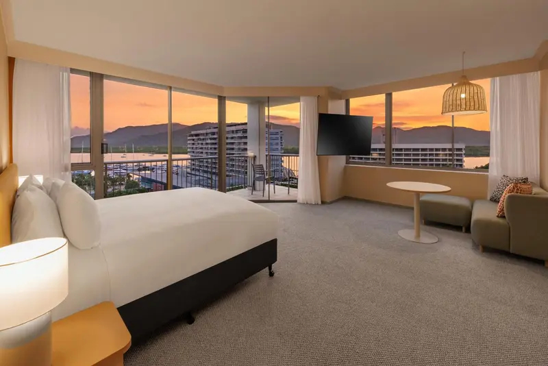 Chambre lumineuse avec un grand lit et une vue sur un paysage urbain au coucher du soleil.