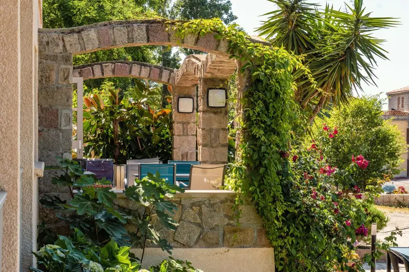Une arche en pierre couverte de végétation donne sur une terrasse aménagée avec des chaises et une table.
