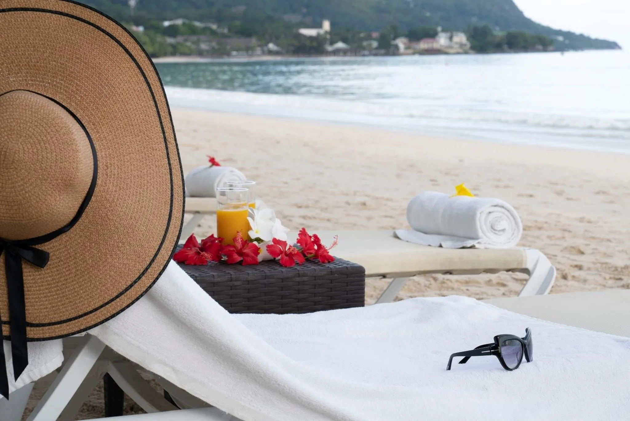 Deux transats blancs sur une plage de sable fin, avec des serviettes, un chapeau et des boissons.