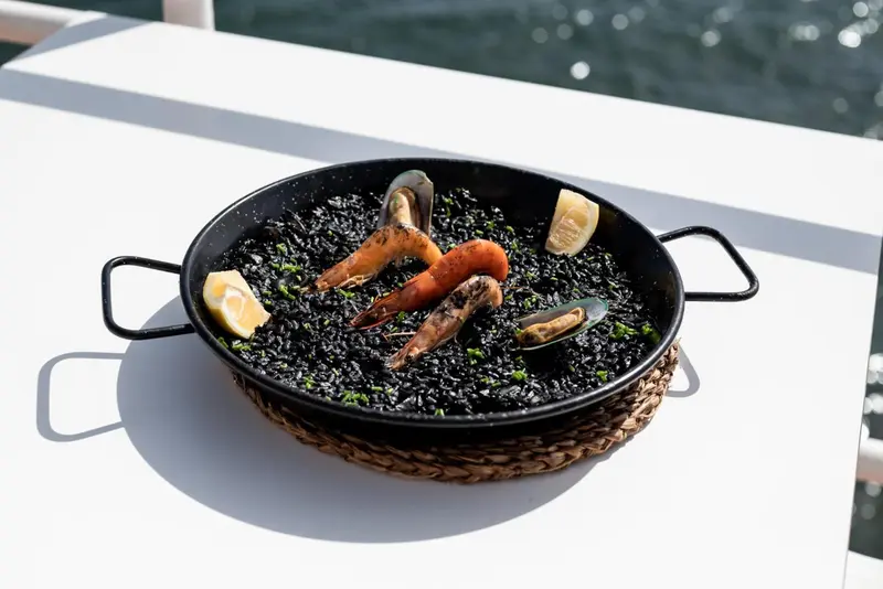 Une grande poêle de paella noire aux fruits de mer est présentée sur une table blanche en extérieur.