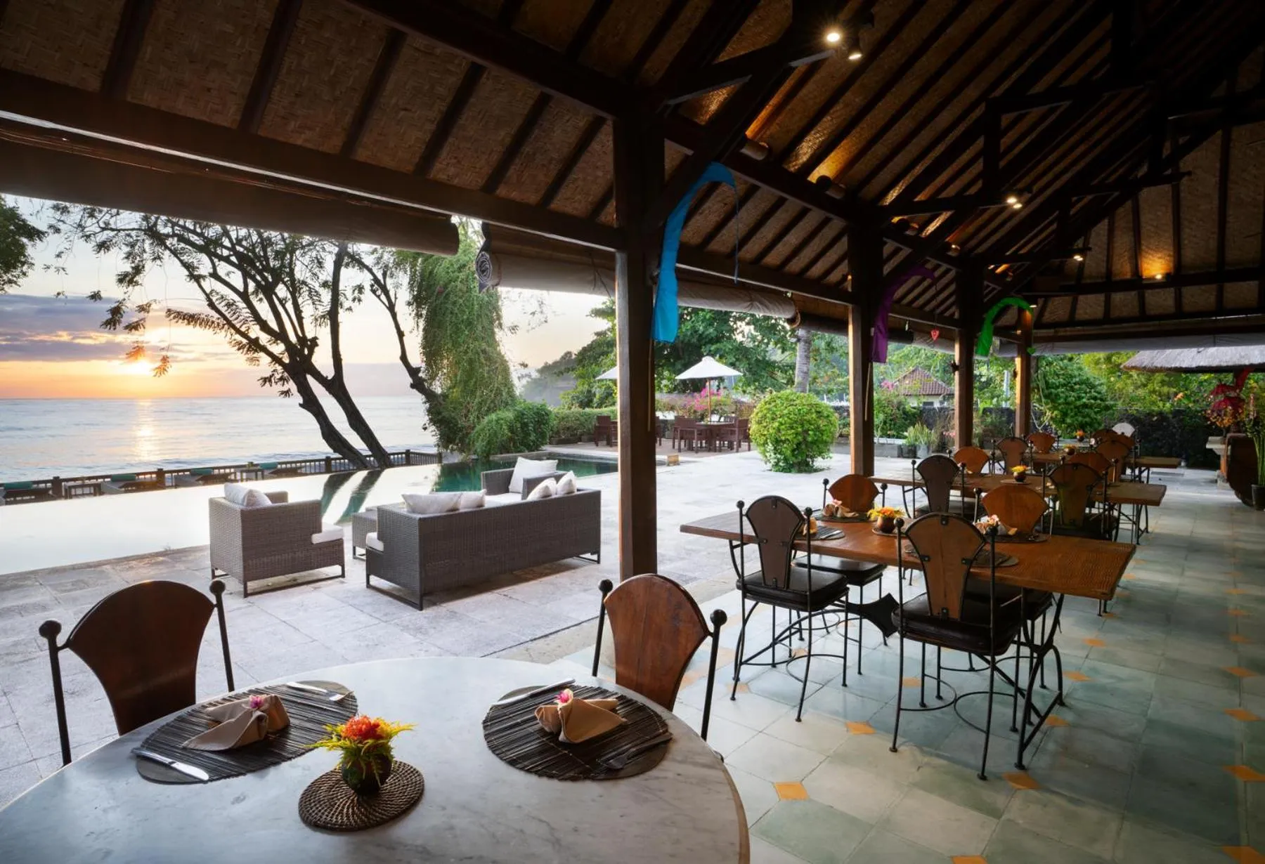 Terrasse ombragée avec tables et canapés face à la mer au coucher du soleil.