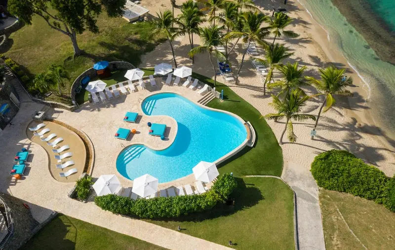 Vue aérienne d'une piscine turquoise bordée de transats sur une plage de sable blanc.