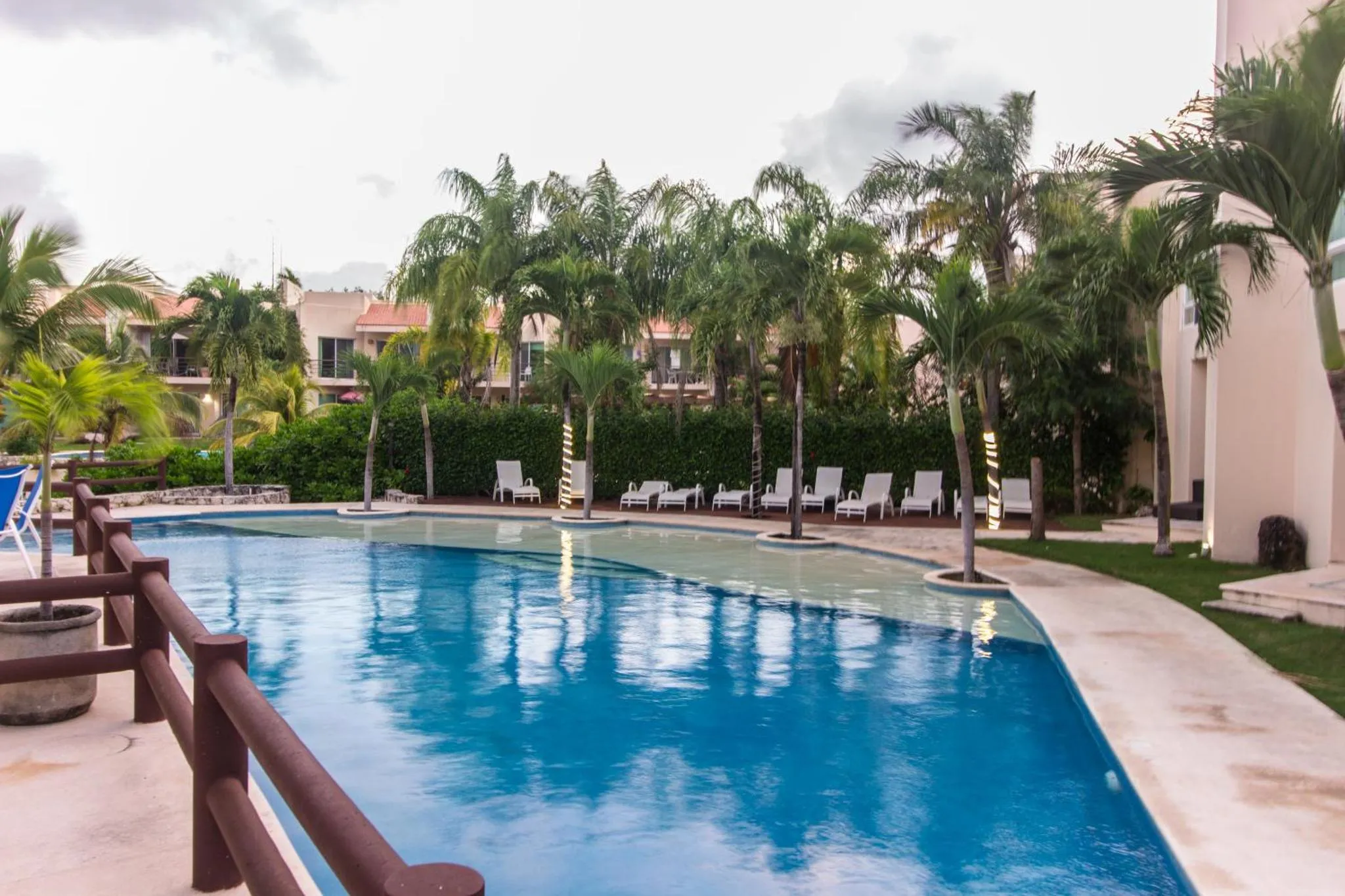 Une piscine bleue bordée de chaises longues, entourée de végétation tropicale et de bâtiments.