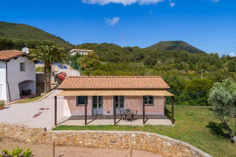Petite maison blanche avec toit en tuiles rouges, entourée de végétation et de collines.