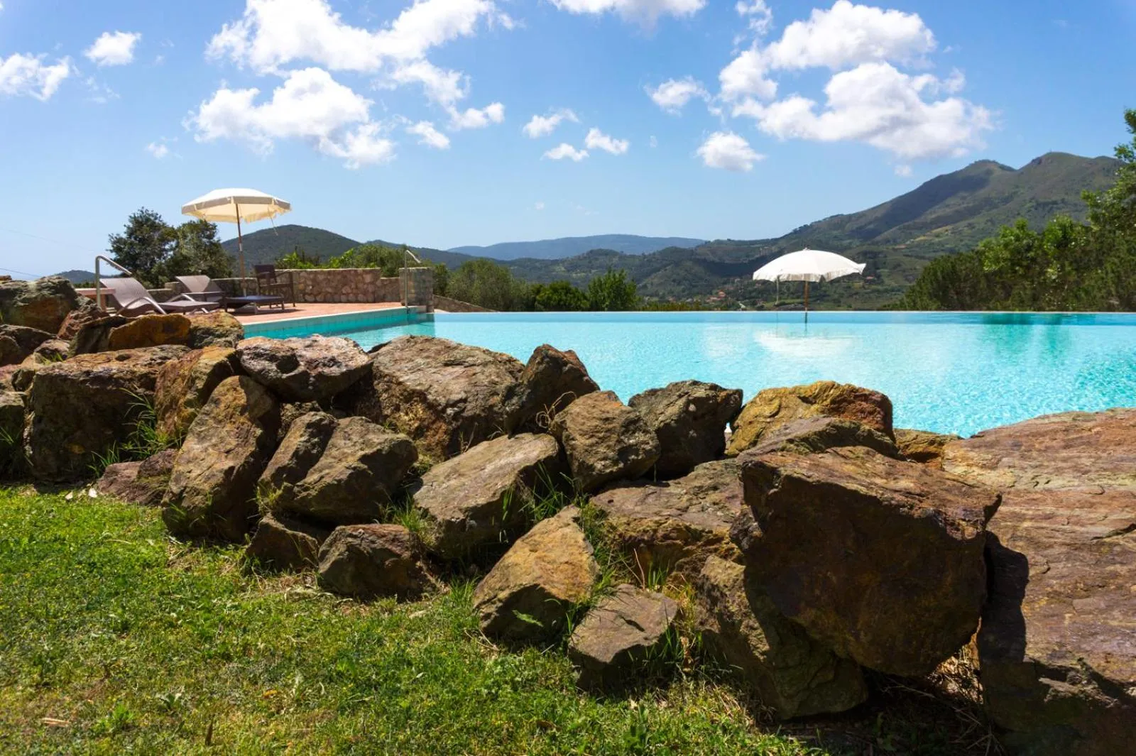 Piscine rectangulaire bordée de rochers et de végétation luxuriante, avec des transats et des parasols.