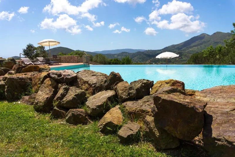 Piscine rectangulaire bordée de rochers et de végétation luxuriante, avec des transats et des parasols.
