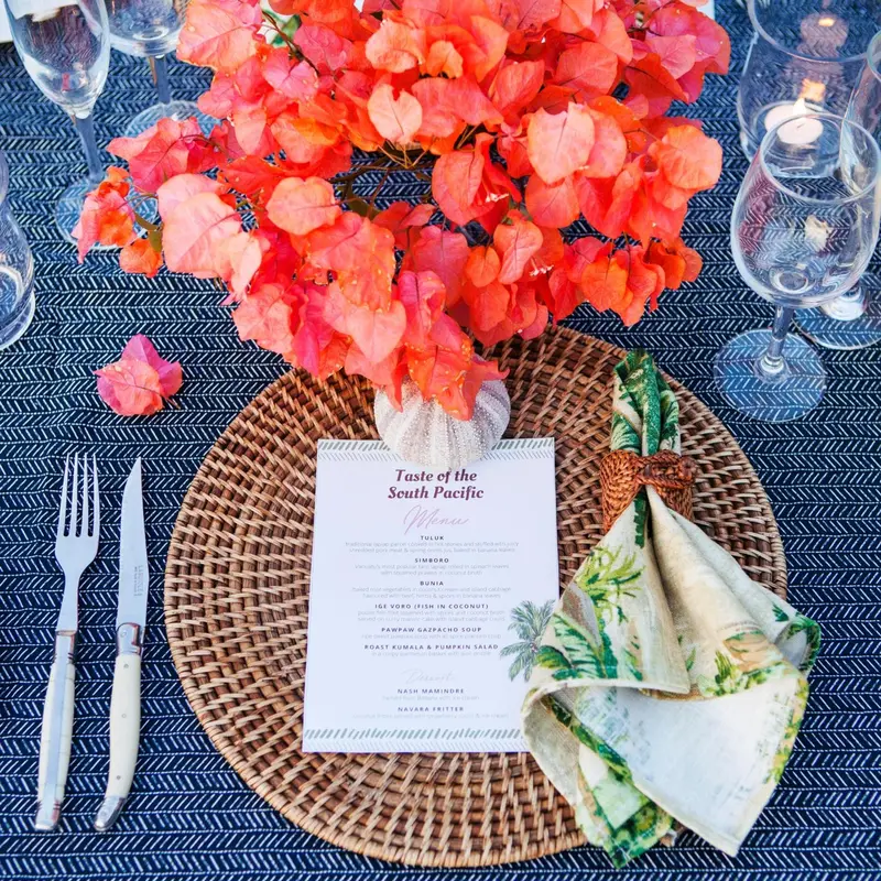 Une table dressée avec une nappe bleue et des sets de table en osier, décorée de fleurs tropicales.