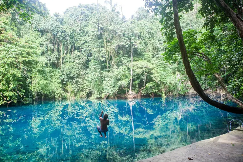 Une personne se balance au-dessus d'une étendue d'eau turquoise bordée de végétation luxuriante.