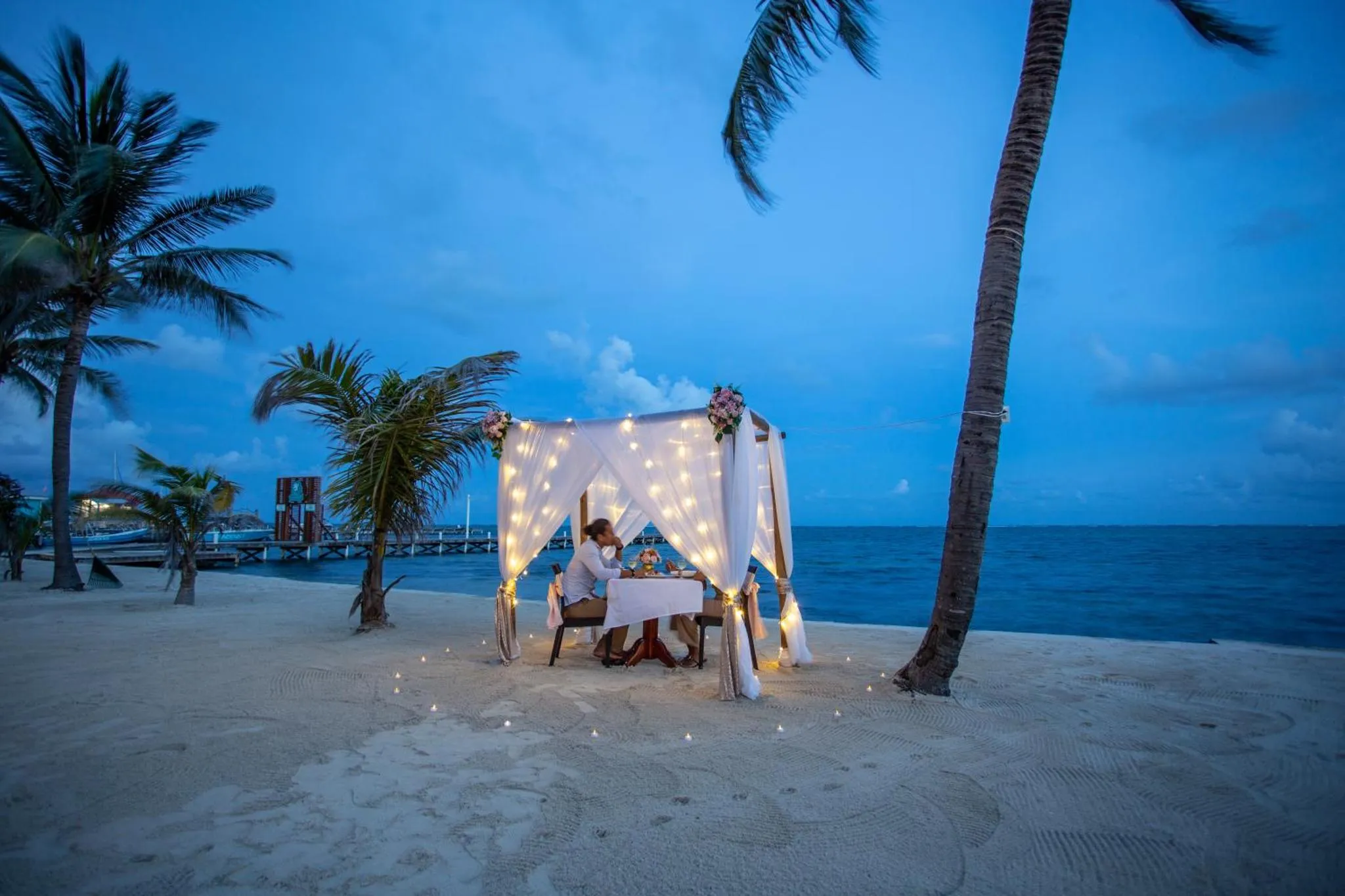 Table dressée pour un dîner romantique sur une plage de sable blanc au coucher du soleil.