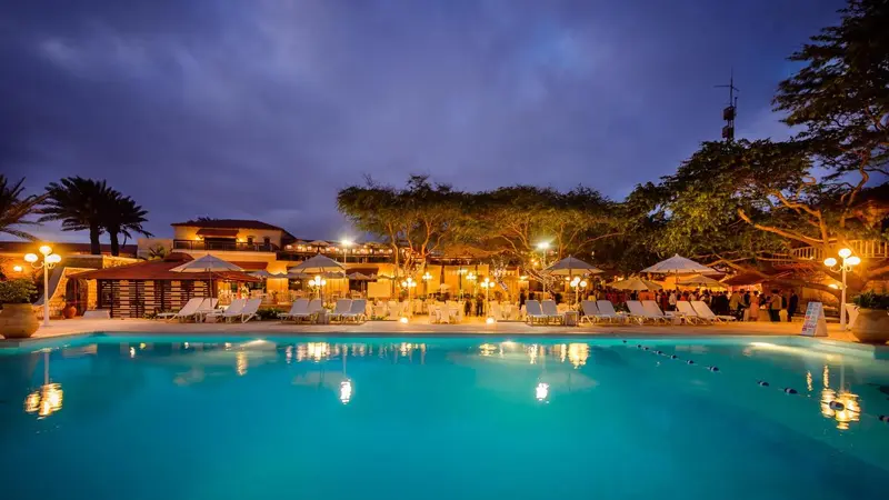 Piscine illuminée devant un bâtiment avec des transats et des palmiers sous un ciel crépusculaire.