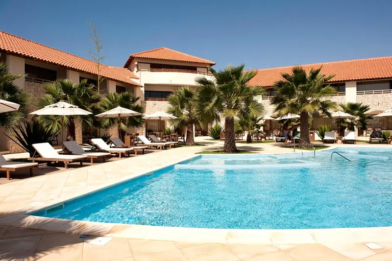 Une piscine turquoise bordée de transats et de parasols devant un bâtiment aux toits de tuiles rouges.