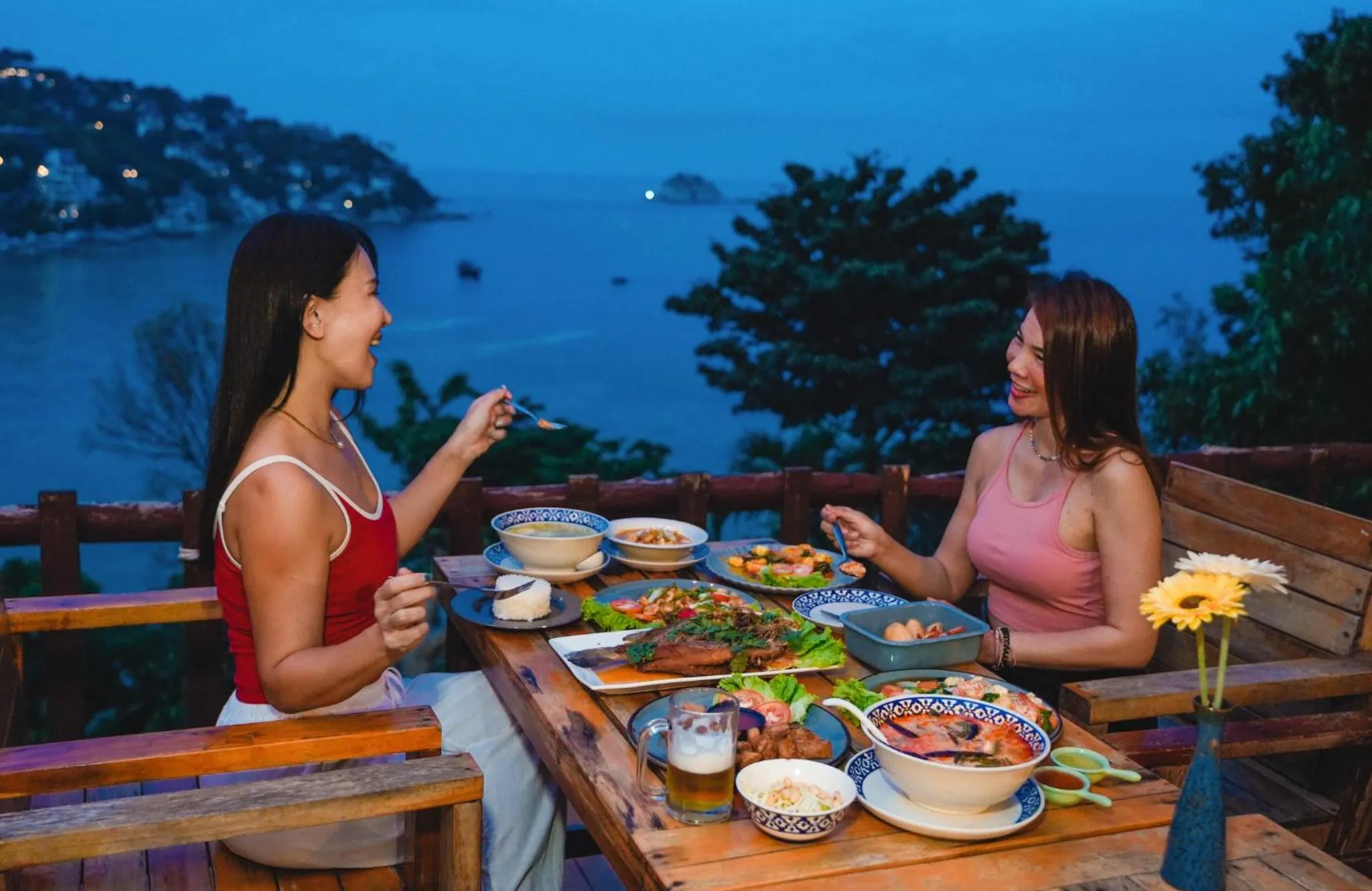 Deux femmes sourient en dégustant un repas sur une terrasse avec vue sur la mer.