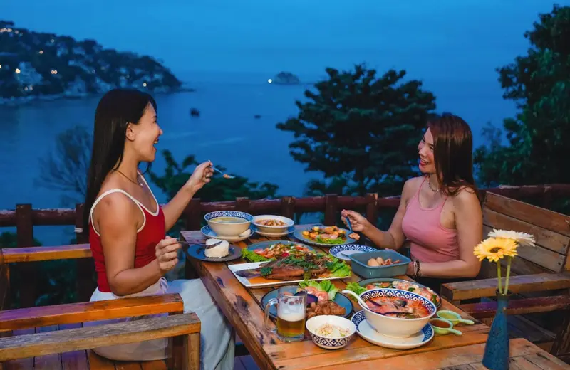 Deux femmes sourient en dégustant un repas sur une terrasse avec vue sur la mer.