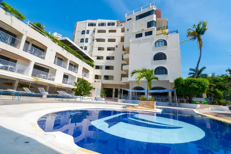 Bâtiment blanc à plusieurs étages avec une piscine au premier plan et des palmiers.