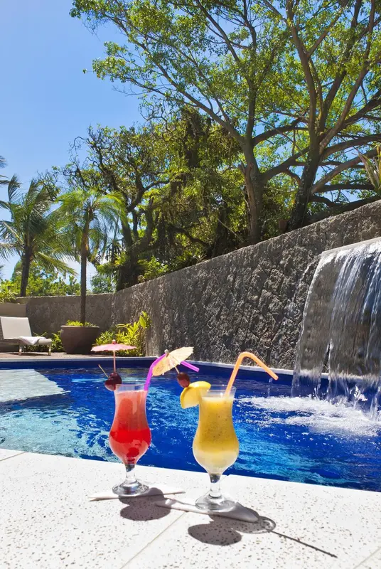 Deux cocktails colorés au bord d'une piscine avec cascade et végétation luxuriante.