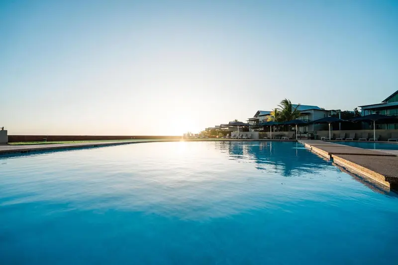 Une longue piscine bleue reflète le soleil couchant devant des bâtiments blancs.