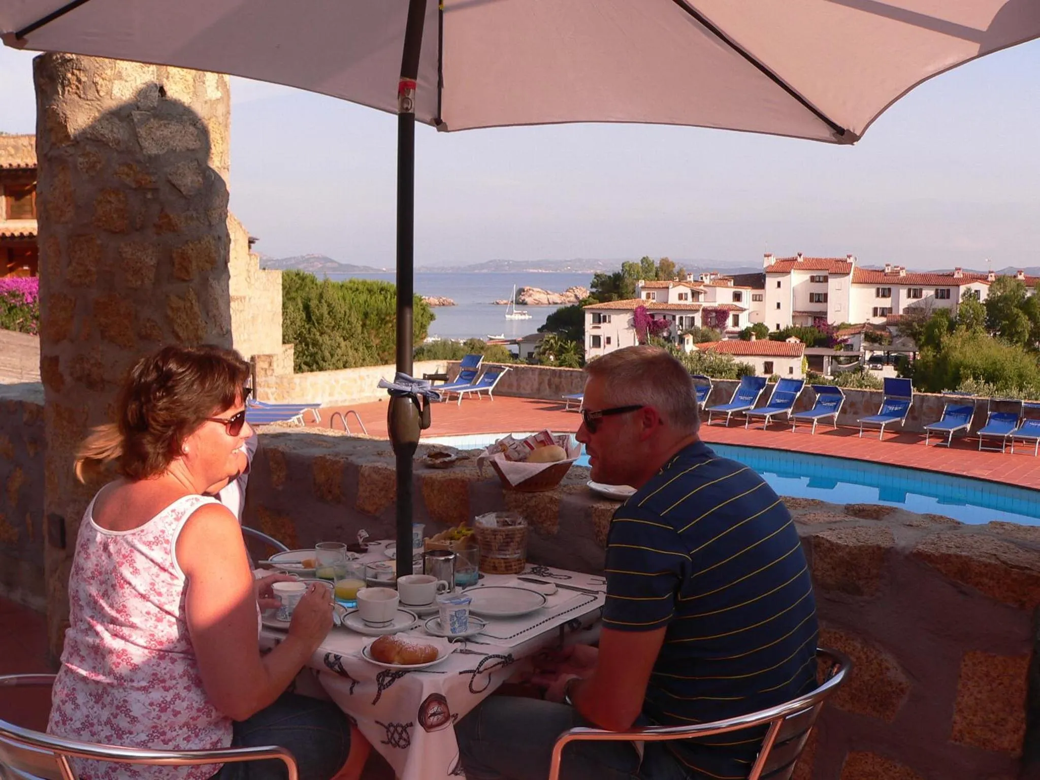 Un couple prend son petit-déjeuner en terrasse avec vue sur la mer et une piscine.