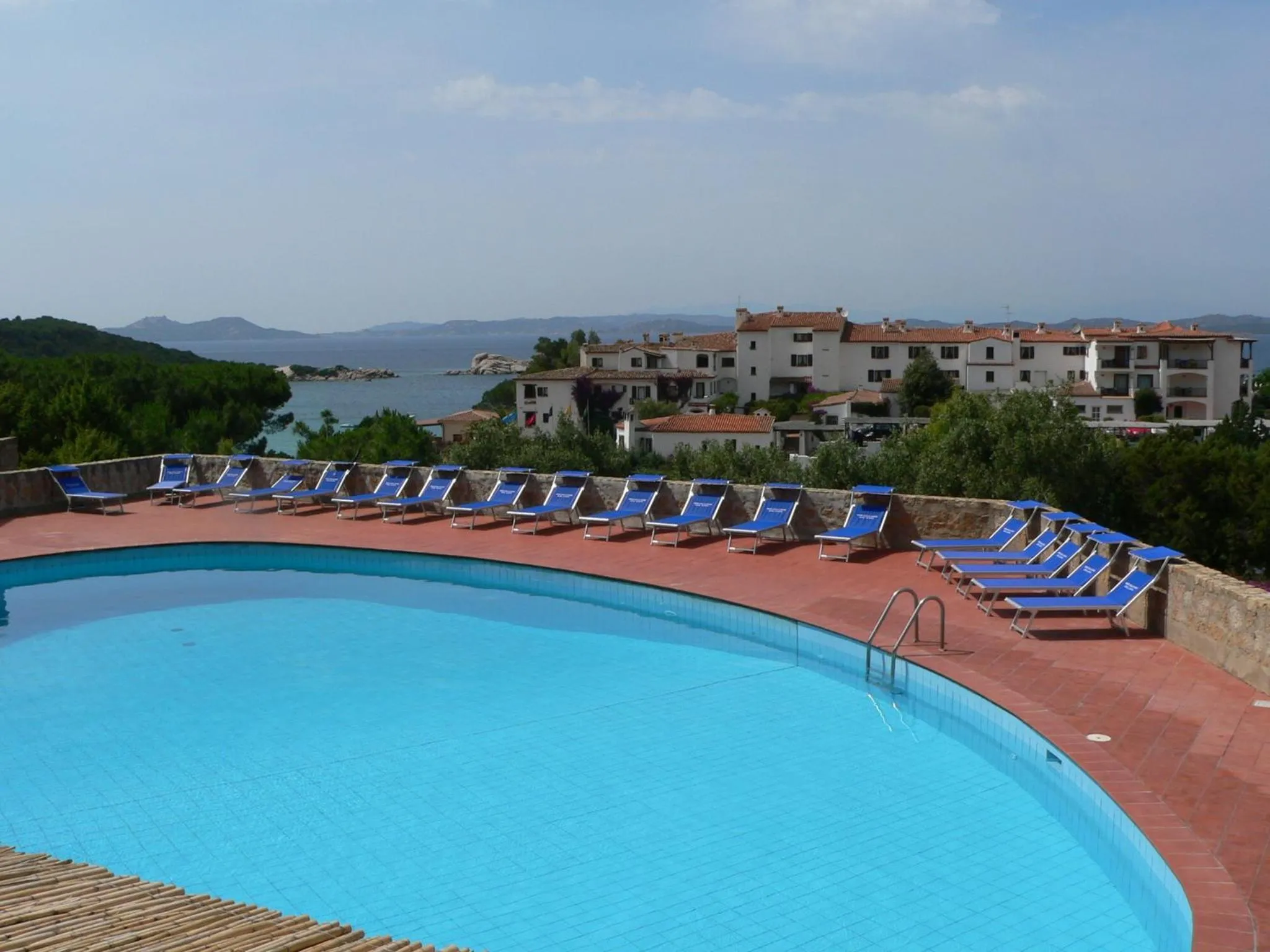 Une piscine turquoise bordée de transats bleus offre une vue sur la mer et les bâtiments environnants.