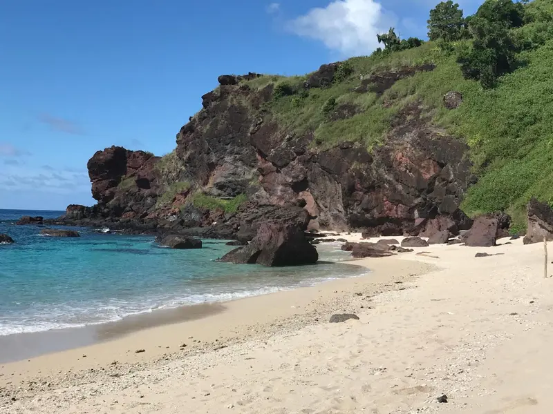 Une plage de sable fin borde une eau turquoise au pied de falaises rocheuses verdoyantes.