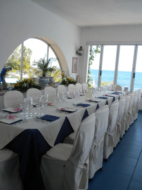 Une longue table est dressée pour un repas dans une salle lumineuse avec vue sur la mer.