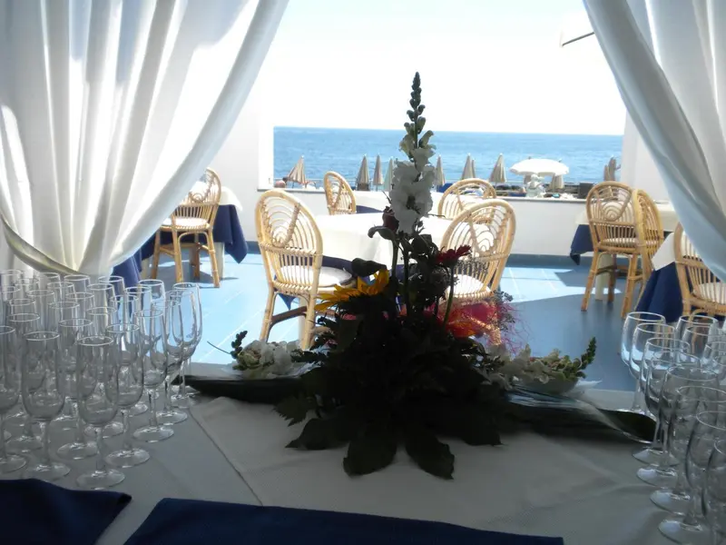 Une table dressée avec des verres et des fleurs en premier plan, donnant sur une terrasse avec des tables et la mer.