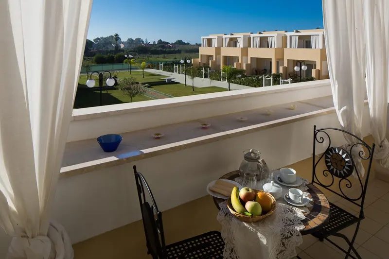 Une table avec un service à thé et des fruits est posée sur un balcon avec vue sur un jardin et des bâtiments.