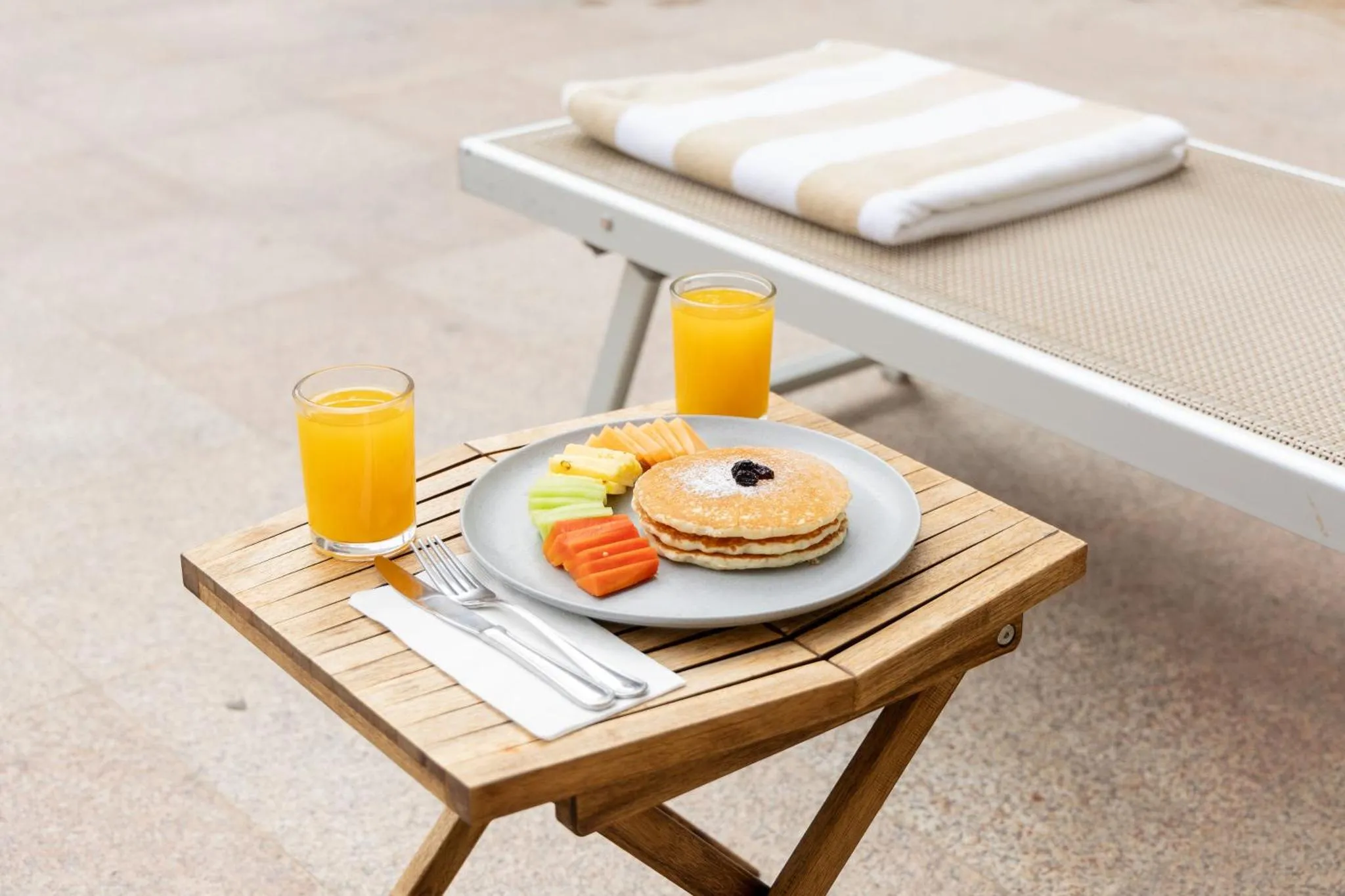 Une table basse en bois présente un petit-déjeuner avec des crêpes, des fruits et des jus d'orange.