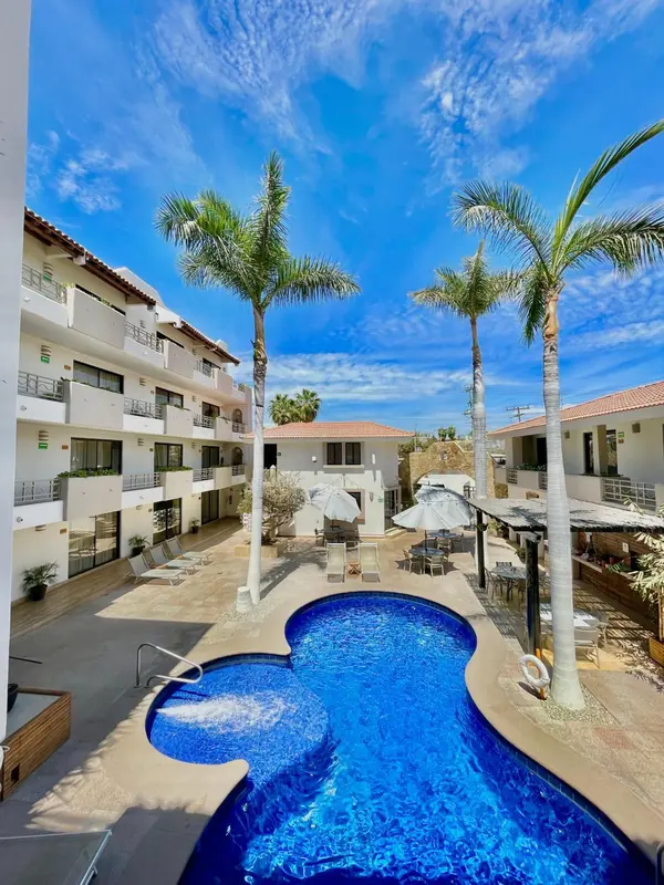 Piscine turquoise au premier plan, bâtiment blanc et palmiers sous un ciel bleu.