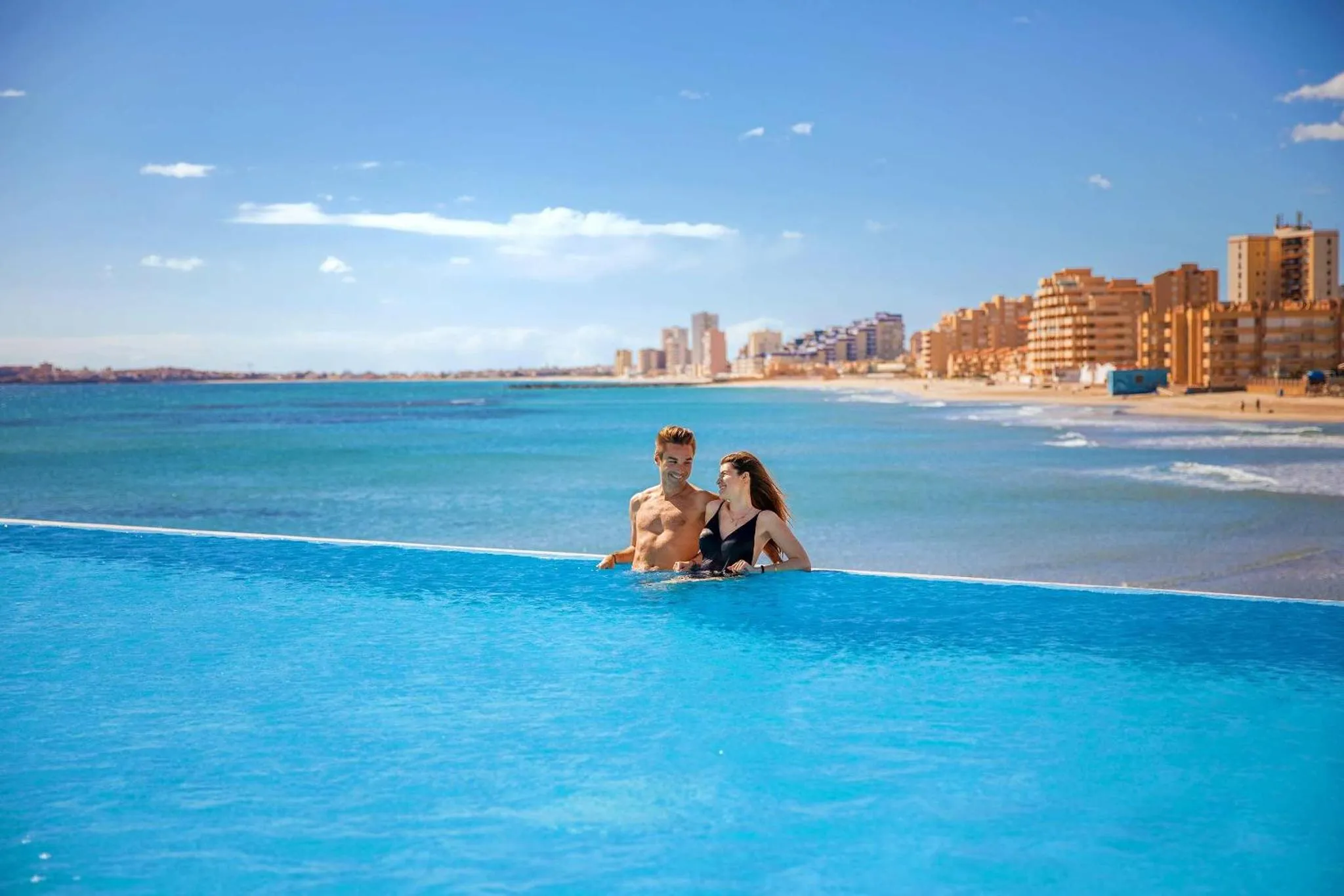 Un couple se tient dans une piscine à débordement avec vue sur la mer et la côte.