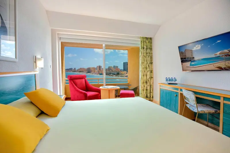 Chambre lumineuse avec un lit et un fauteuil face à une grande fenêtre donnant sur la mer.