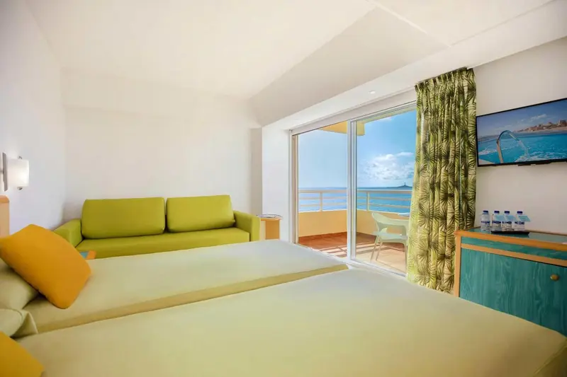 Une chambre lumineuse avec un canapé vert et une vue sur la mer depuis un balcon.