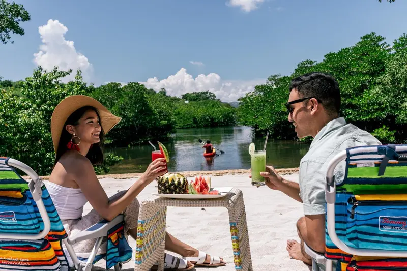 Un couple souriant déguste des boissons et des fruits sur une plage ensoleillée.