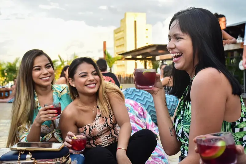 Trois jeunes femmes sourient et trinquent avec des cocktails sur une terrasse ensoleillée.