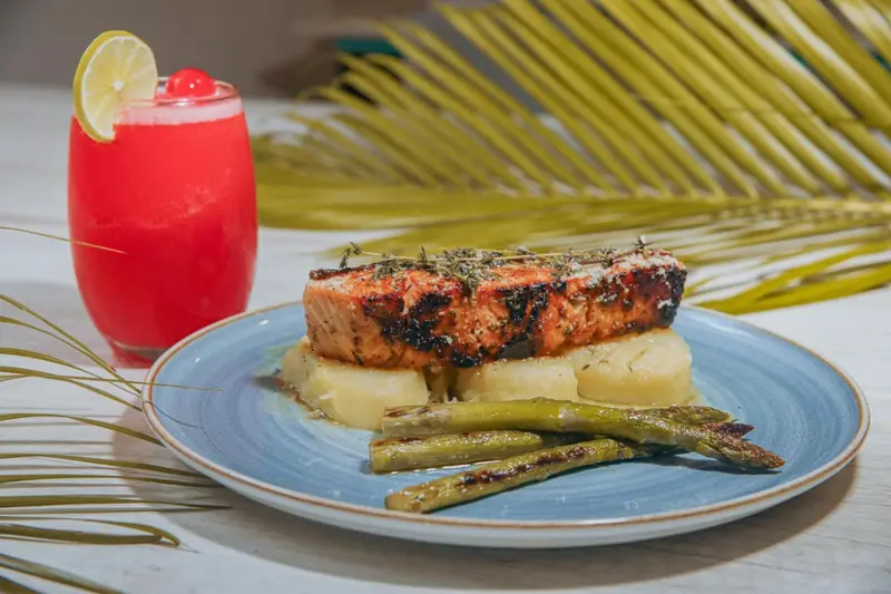 Un plat de poisson grillé accompagné de légumes et d'une boisson rose est présenté sur une table.
