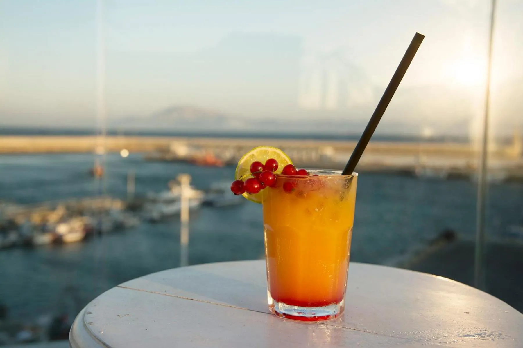Un cocktail coloré est présenté sur une table blanche avec vue sur la mer et un port.