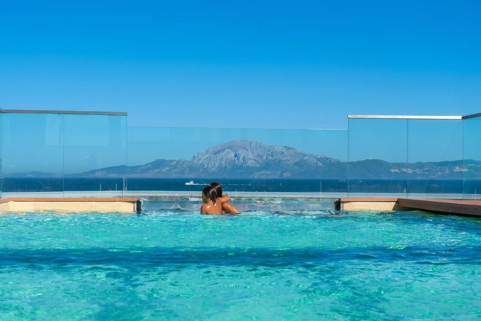 Deux personnes se détendent dans une piscine à débordement avec vue sur la mer et les montagnes.