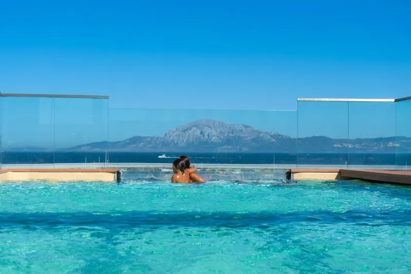 Deux personnes se détendent dans une piscine à débordement avec vue sur la mer et les montagnes.