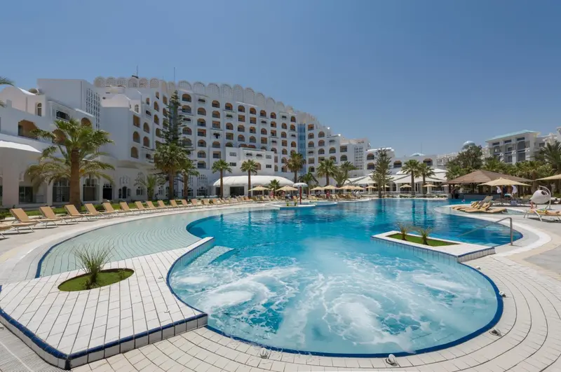 Une grande piscine turquoise avec un jacuzzi intégré, entourée de chaises longues et de palmiers devant un bâtiment blanc.