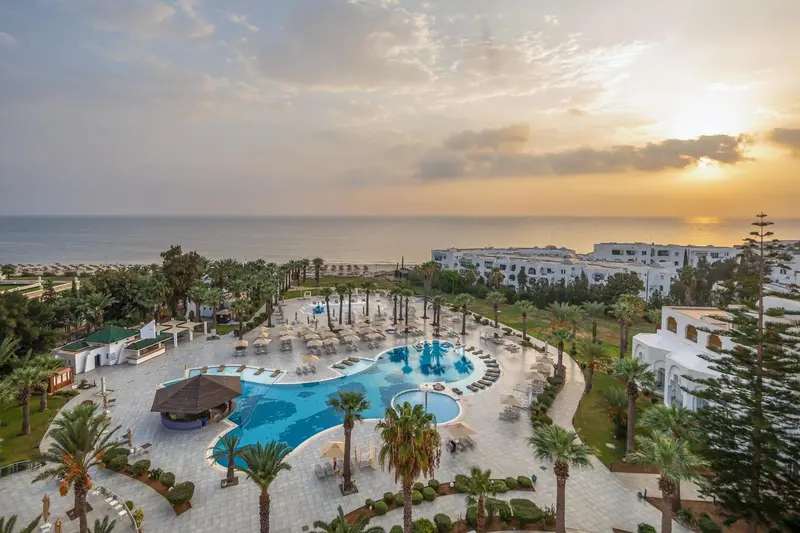 Vue aérienne d'un complexe hôtelier avec piscine et palmiers au coucher du soleil.