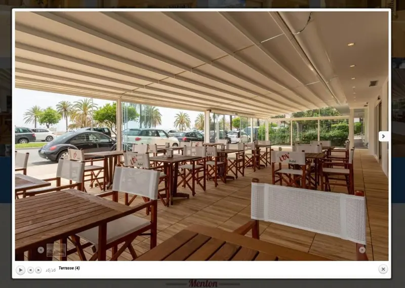 Terrasse ombragée avec tables et chaises donnant sur un parking et des palmiers.