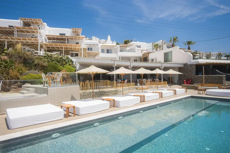 Transats blancs alignés au bord d'une piscine turquoise avec en arrière-plan des bâtiments blancs et des parasols.