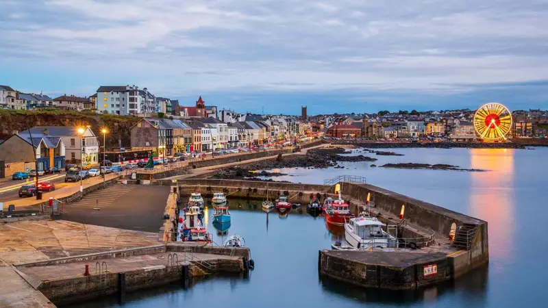 Vue panoramique d'un port de ville au crépuscule, avec des bateaux amarrés et des bâtiments illuminés.