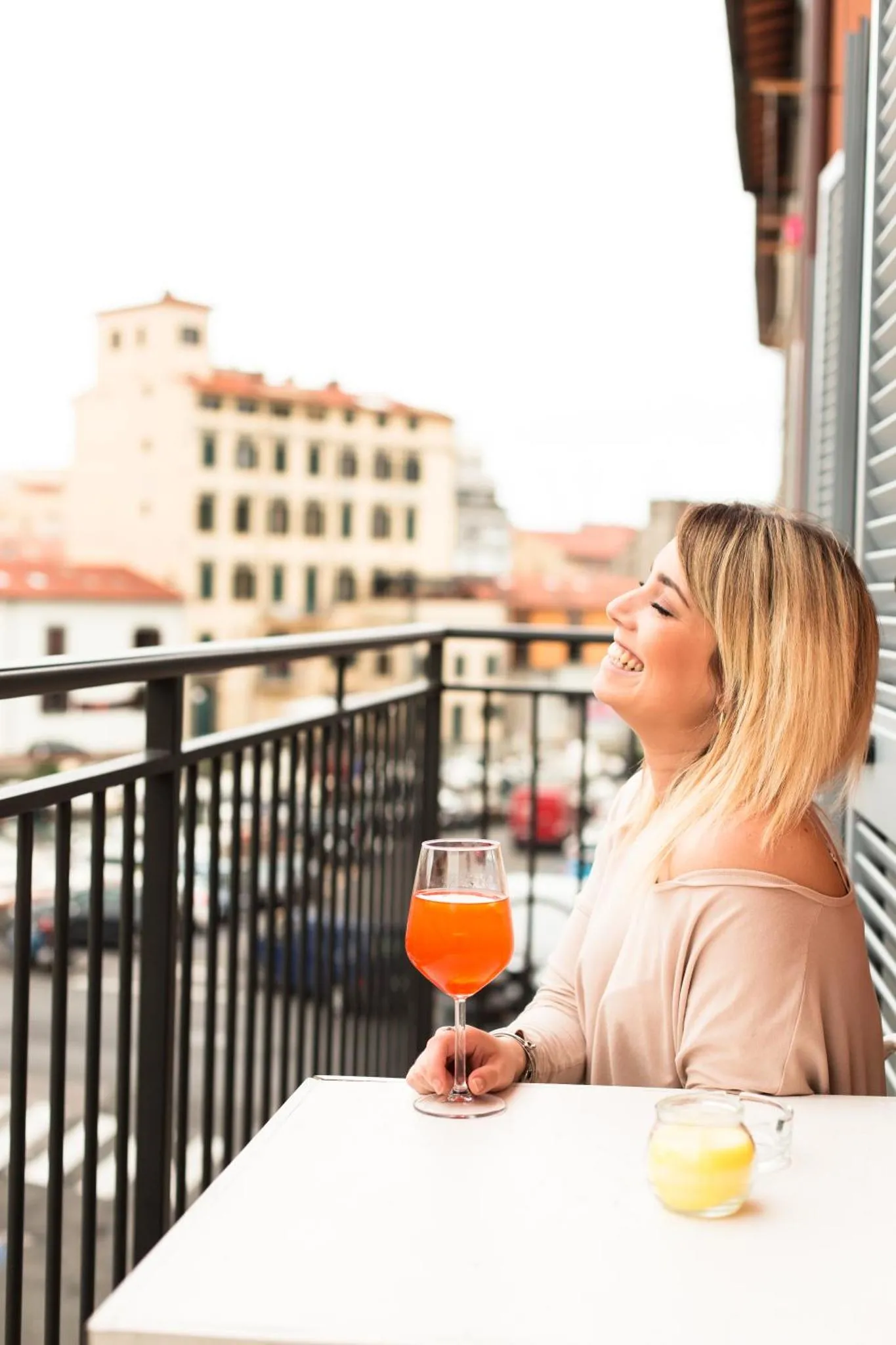 Une femme souriante tient un verre sur une terrasse ensoleillée avec vue sur la ville.