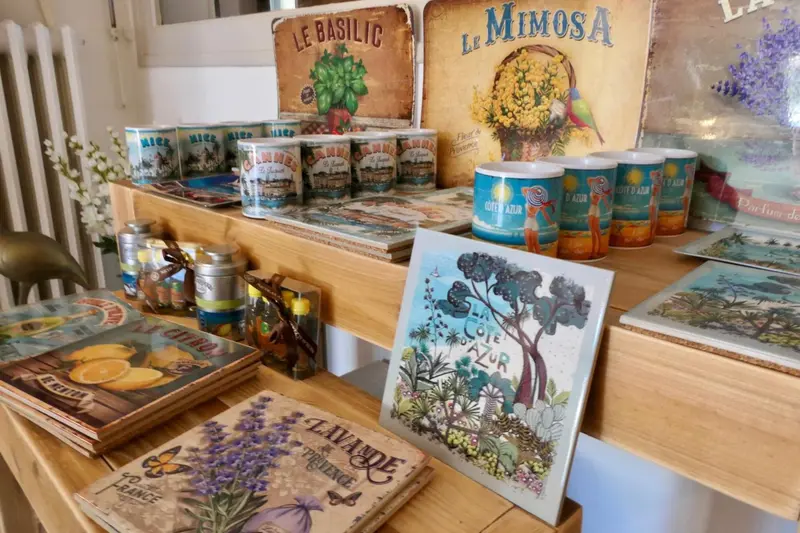 Une étagère en bois présente des tasses, des toiles et des tableaux décoratifs aux motifs floraux et provençaux.