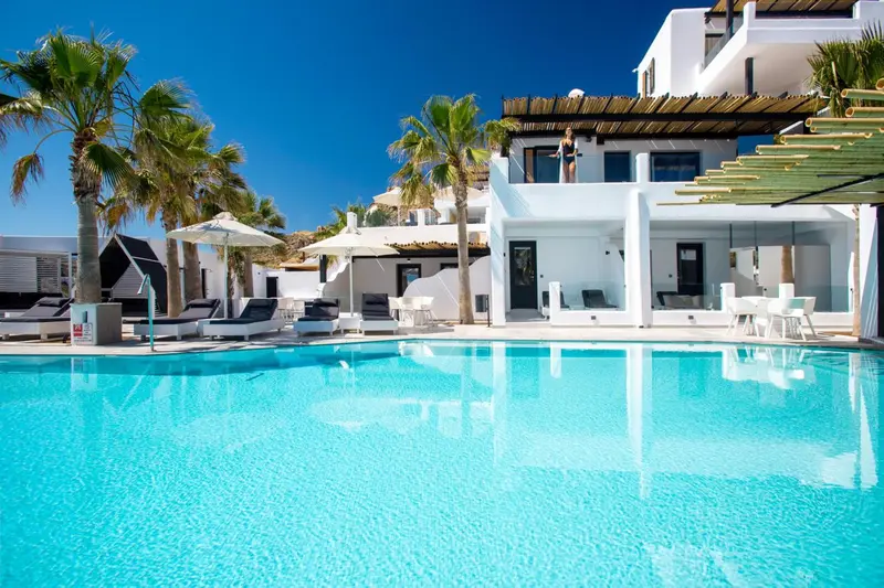 Une piscine turquoise reflète un bâtiment blanc et des palmiers sous un ciel bleu.