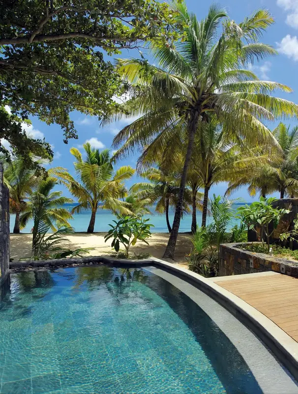 Une piscine turquoise bordée d'une terrasse en bois, avec des palmiers et une plage en arrière-plan.