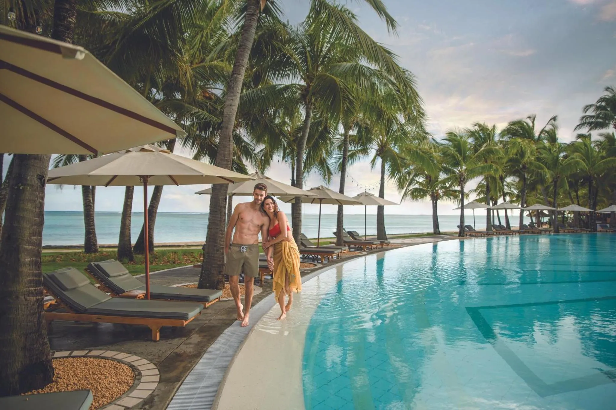 Un couple se promène au bord d'une piscine turquoise bordée de transats.