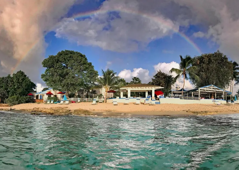 Plage de sable fin bordée de palmiers et d'une végétation luxuriante sous un ciel nuageux illuminé d'un arc-en-ciel.