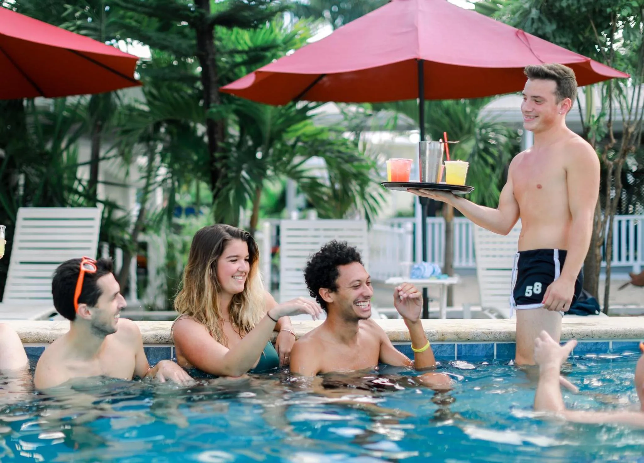 Des clients sourient et discutent au bord d'une piscine ensoleillée, tandis qu'un serveur leur apporte des boissons.