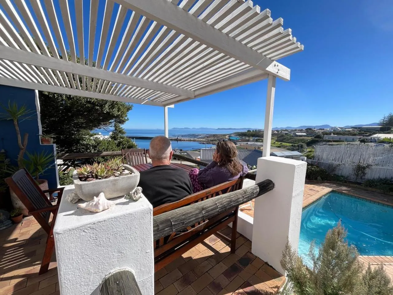 Deux personnes sont assises sur un banc face à la mer sous une pergola.